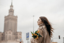 A Young Girl In Glasses With Flowers Against The Background Of The Palace Of Culture In Warsaw. A Pretty Girl In Glasses With Flowers Walks In Warsaw.