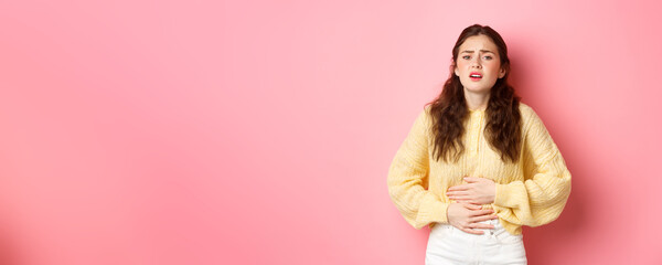 Wall Mural - Young woman with pain in stomach, holding hands on belly, feeling terrible ache, menstrual cramps, standing against pink background