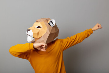 Horizontal shot of strong man with lion mask with wearing casual style orange jumper posing isolated over gray background, standing with raised arms and clenched fists, looking away.