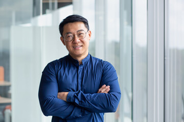 Portrait of successful mature boss, senior businessman in glasses Asian looking at camera and smiling, man with crossed arms working inside modern office building.