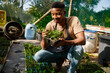 © StratfordProductions - Happy young black man wearing apron and examining the plants at garden center