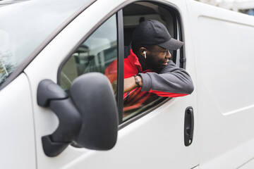 Wall Mural - Young adult black delivery guy wearing work unifrom driving white van leaning out the window to look behind. Horizontal shot. High quality photo