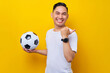 © Bangun Stock Photo - Excited young Asian man football fan wearing a white t-shirt holding a soccer ball and pointing his thumb in a blank space isolated on yellow background. People sport leisure lifestyle concept