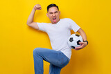 Excited young Asian man 20s wearing a white t-shirt supporting a football sports team, holding in hand a soccer ball and watching tv live stream do winner gesture isolated on yellow background