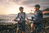 Senior caucasian couple riding off road on the pebble beach with electric bicycles at sunset. Authentic elderly retired life and sustainable mobility concept