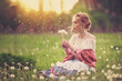 Beautiful girl in a vintage look sits at sunset in a field of dandelions and blows on dandelions, spring