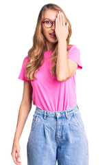 Wall Mural - Young beautiful caucasian woman with blond hair wearing casual clothes and glasses covering one eye with hand, confident smile on face and surprise emotion.