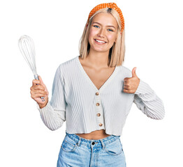 Canvas Print - Beautiful young blonde woman holding baker whisk smiling happy and positive, thumb up doing excellent and approval sign