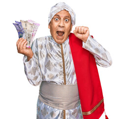 Poster - Senior hispanic man wearing sherwani costume holding indian rupee banknotes screaming proud, celebrating victory and success very excited with raised arms