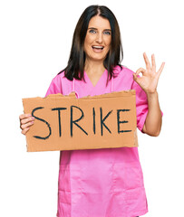 Canvas Print - Middle age brunette doctor woman holding strike banner cardboard doing ok sign with fingers, smiling friendly gesturing excellent symbol