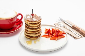 Canvas Print - Honey pancakes with a sliced strawberry on a white plate, with a cup of coffee and cutlery