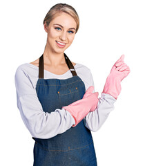 Wall Mural - Beautiful young blonde woman wearing cleaner apron and gloves smiling happy pointing with hand and finger to the side