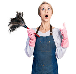 Poster - Beautiful young blonde woman wearing apron holding cleaning duster amazed and surprised looking up and pointing with fingers and raised arms.