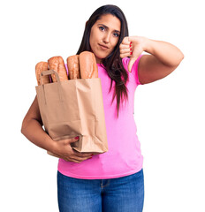 Sticker - Young beautiful brunette woman holding delivery bag with bread with angry face, negative sign showing dislike with thumbs down, rejection concept