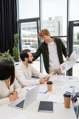 Wall Mural - smiling man with infographics talking to interracial colleagues in meeting room