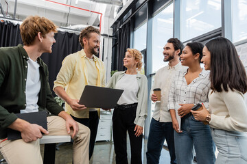 Wall Mural - cheerful man with laptop talking to smiling interracial business colleagues in office