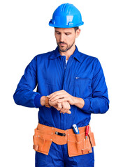 Canvas Print - Young handsome man wearing worker uniform and hardhat in hurry pointing to watch time, impatience, upset and angry for deadline delay