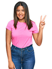 Wall Mural - Young latin girl wearing casual clothes smiling with happy face winking at the camera doing victory sign. number two.