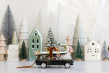 Holidays Are Coming! Stylish Little Car Carrying Candy Cane On Background Of Christmas Miniature Snowy Village. Merry Christmas! Festive Winter Scene On White Table, Xmas Banner