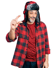 Sticker - Middle age hispanic man with grey hair wearing fluffy earmuff hat smiling and confident gesturing with hand doing small size sign with fingers looking and the camera. measure concept.