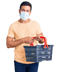 Sticker - Young hispanic man wearing shopping basket and medical mask thinking attitude and sober expression looking self confident