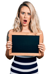 Poster - Young blonde woman holding blackboard in shock face, looking skeptical and sarcastic, surprised with open mouth