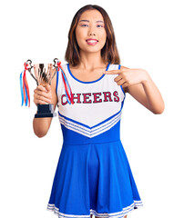 Youn beautiful asian girl wearing cheerleader uniform holding champion trophy smiling happy pointing with hand and finger