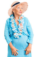 Senior beautiful woman with blue eyes and grey hair wearing summer hat and hawaiian lei looking away to side with smile on face, natural expression. laughing confident.