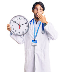 Poster - Handsome hispanic man wearing doctor uniform holding clock covering mouth with hand, shocked and afraid for mistake. surprised expression