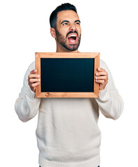 Canvas Print - Young hispanic man with beard holding blackboard angry and mad screaming frustrated and furious, shouting with anger. rage and aggressive concept.