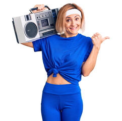 Sticker - Young blonde woman wearing sportswear holding boombox, listening to music pointing thumb up to the side smiling happy with open mouth