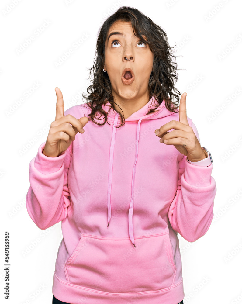 Young hispanic woman wearing casual sweatshirt amazed and surprised looking up and pointing with fingers and raised arms.