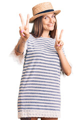 Wall Mural - Beautiful caucasian woman wearing summer hat smiling looking to the camera showing fingers doing victory sign. number two.