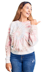 Sticker - Beautiful young woman wearing casual tie dye sweatshirt smiling with happy face looking and pointing to the side with thumb up.