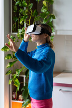 Young Woman Wearing VR Glasses Plays Futuristic Game With Amazing Graphics At Home. Female Wearing Blue Sweater Explores Virtual World Of Game Standing Against High Green Houseplants At Daylight
