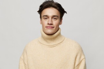 Horizontal studio shot. A handsome young man with dark short hair combed back in a beige sweater with a high neck stands on a gray background