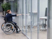Red-haired Caucasian Woman In A Wheelchair Trying To Open The Door In The Office. 