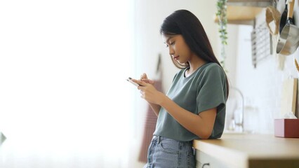 Poster - Happy young asian woman relaxing at home. Asia female standing at counter kitchen and using mobile smartphone