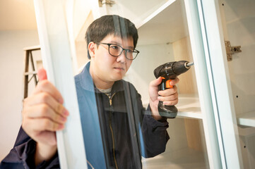 Wall Mural - Asian male furniture assembler using electric drill screwdriver on cabinet hinge of glass panel. Interior construction worker man using screwdriving tools for furniture installation in the new house.