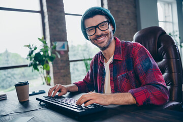 Photo of happy smiling coder dressed hat glasses typing program modern gadget indoors workplace workstation loft