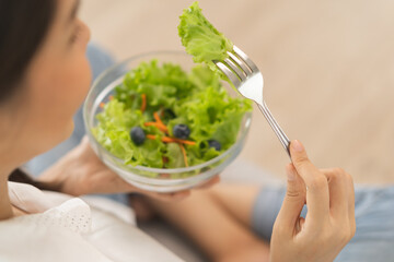 Wall Mural - Diet, Dieting asian young woman, girl hand use fork at lettuce on mix vegetables, green salad bowl, eat food low fat to good body sitting on sofa at home. Nutritionist female, Weight loss for healthy.