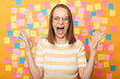 © sementsova321 - Image of angry aggressive woman with brown hair in striped t shirt standing against yellow wall with colorful stickers, raised arms and yelling with hate.