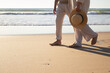 Couple enjoying vacation, walking along seashore with seascape in background. Legs closeup of lady in cardigan with straw hat and man strolling barefoot on wet sand. Romance, holiday concept