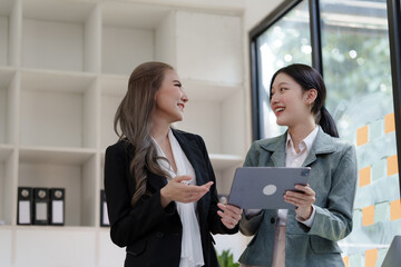 Pretty Asian woman in black business suit consults with her colleague, Two business women discuss working issues in office using documents and tablet.