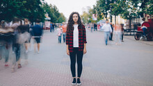 Portrait Of Beautiful Young Girl With Long Curly Hair Standing In Street Looking At Camera When Many Men And Women Are Walking Around In Hurry On Summer Day.