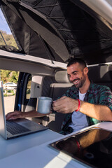 smiling young man making a video call with his laptop sitting in his camper van, concept of freedom and digital nomad lifestyle