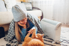 Using Heater At Home In Winter. Woman Playing With Cat By Device Wearing Sweater And Hat Under Blanket. Heating Season.