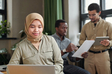 Poster - Young Muslim businesswoman in hijab and casual attire sitting in front of laptop and networking against two intercultural male coworkers