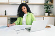 © shurkin_son - Female lawyer freelancer with dark skin and curly hair, working from home sitting at kitchen table in front of laptop, writing in her copybook, looking at camera smiling, dressed in light green shirt