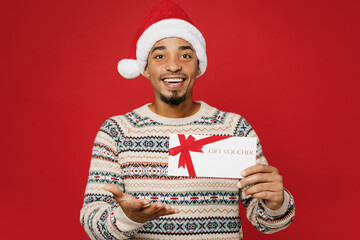 Sticker - Merry young man wear cozy Christmas sweater Santa hat posing hold give gift certificate coupon voucher card for store isolated on plain red background. Happy New Year 2023 celebration holiday concept.